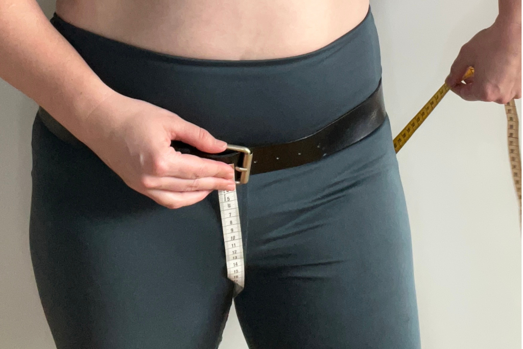 A photo of a model holding a measuring tape against the buckle of a belt while the other hand holds the tape which travels between their legs and behind them