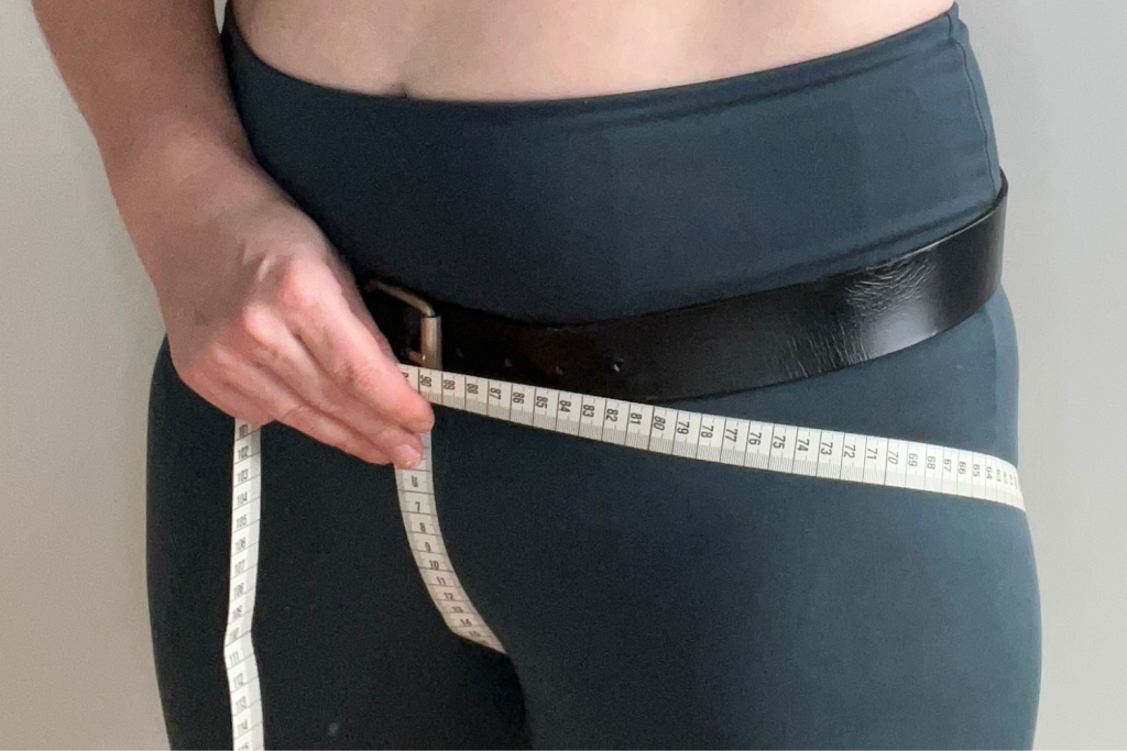 A photo of a model holding both ends of a measuring tape against the buckle of a belt sitting low on their hips
