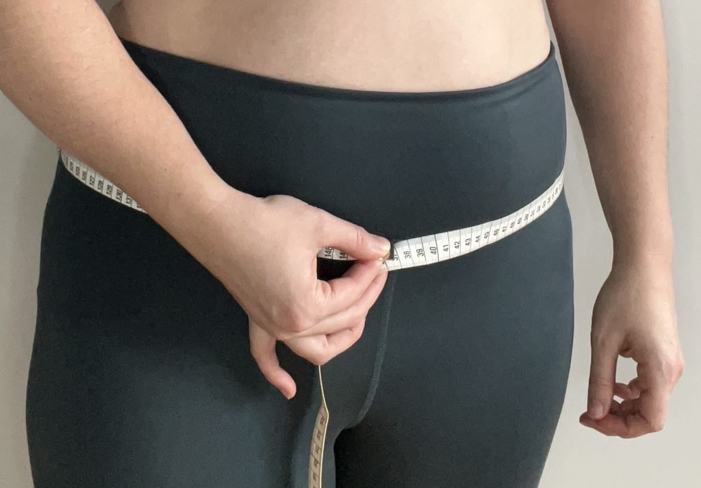 A close up photograph of the hip area of a model wearing dark leggings. In one hand they hold a measuring tape which is wrapped around their hips.